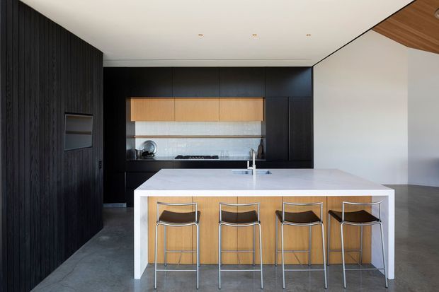 A sleek contemporary kitchen with a large white island and four bar stools. The kitchen features extensive dark vertical timber cabinetry, light timber overhead cabinetry, a pale stone-look benchtop and splashback, integrated appliances, and a polished concrete floor. The space has a clean, minimalist composition with a partly timber-lined angled ceiling.
