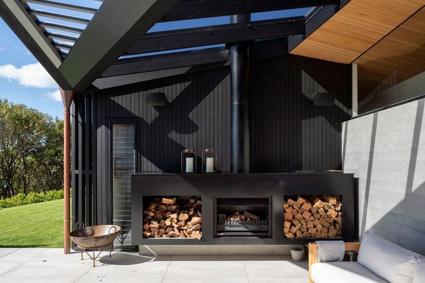 Contemporary outdoor entertaining area with a built-in dark masonry fireplace and central firebox, flanked by open firewood storage niches. The space features black vertical wall cladding, exposed dark structural beams, a timber-lined ceiling and roof structure, concrete paving, and a partially enclosed seating area overlooking landscaped grounds.