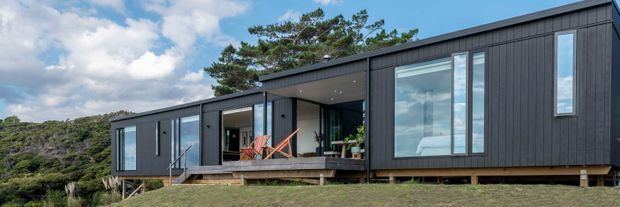 A modern single-storey modular-style residence set in a natural, elevated landscape. The elongated building has dark vertical cladding, a low-pitched roof, expansive floor-to-ceiling glazing, and a recessed covered outdoor terrace with timber decking and seating. The structure appears raised slightly above the ground on supports, with rocky vegetation and trees surrounding it.