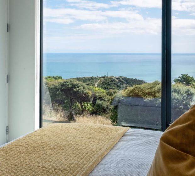 A bedroom interior with a bed positioned beside expansive floor-to-ceiling glazing, looking out over coastal vegetation and a blue ocean horizon. The room has a simple white wall and dark window frame, with a textured mustard quilt and warm brown bedding creating a calm, natural coastal atmosphere.