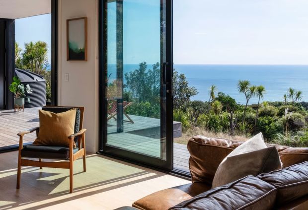 A bright coastal living room opening onto a deck with expansive ocean views. The room features full-height black-framed glazed doors, pale timber flooring, a brown leather sofa, a mid-century-style wooden armchair with tan cushions, and a framed wall artwork. Native coastal planting and the sea are visible beyond the glazing.