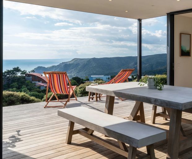 A bright contemporary outdoor dining area on a timber deck, with a large concrete-look dining table and bench in the foreground, colourful striped deck chairs, and expansive ocean and mountainous coastal views beyond. The space is partially sheltered by a flat roof with slim dark structural posts and opens directly to the landscape.