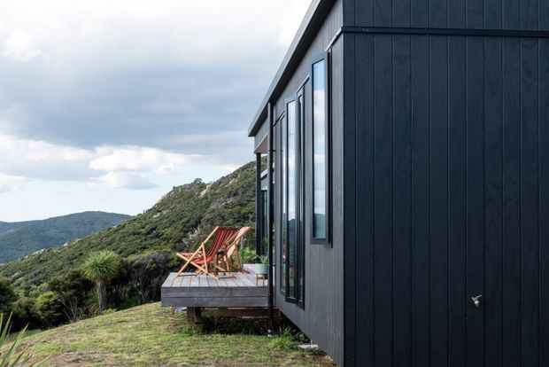Exterior side view of a contemporary dark-clad building on a steep, elevated bush-covered site. The facade has vertical dark timber-like cladding, tall narrow windows, and a projecting timber deck with outdoor chairs, overlooking mountainous terrain beneath a cloudy sky.