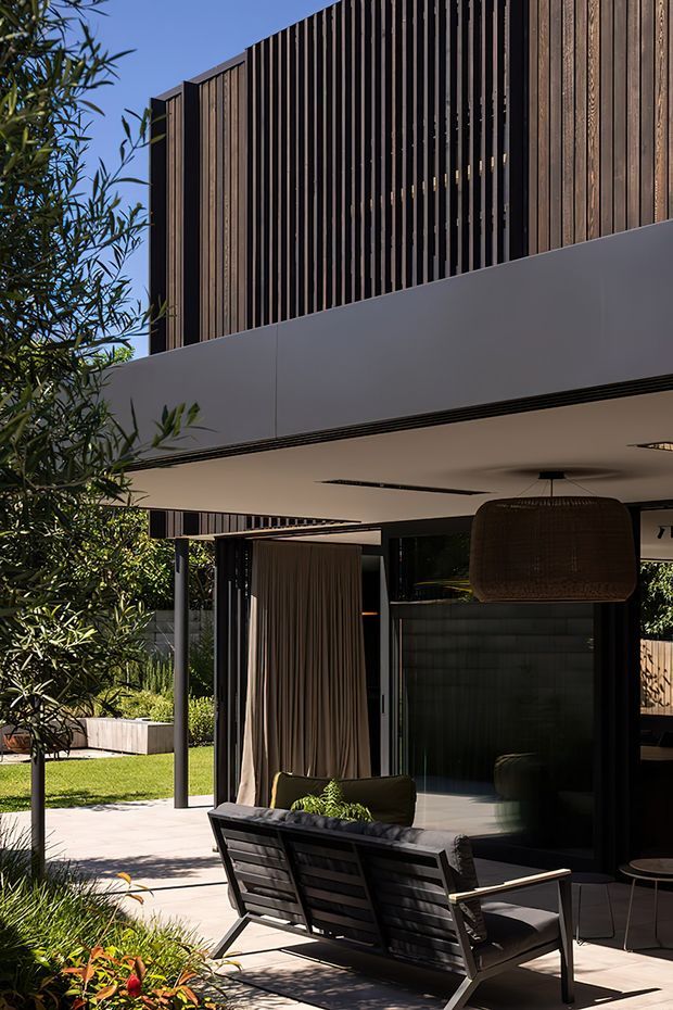 A contemporary multi-level residential exterior with a covered outdoor entertaining area. The facade combines dark vertical timber screening, black-framed glazing, and smooth light-grey panels. A lounge setting with an outdoor sofa, suspended woven pendant light, and curtains sits beneath the cantilevered upper level, opening onto a landscaped garden and paved terrace.