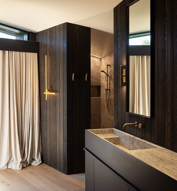 A moody, contemporary bathroom with dark vertically grained timber wall panelling and joinery, a large black-framed mirror, brass tapware and handles, a stone vanity benchtop with integrated basin, and a tiled walk-in shower. Full-height cream curtains and pale timber flooring are visible at the left.