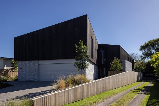 Contemporary multi-level residential building viewed from the street, featuring dark vertical timber-clad upper volumes, pale masonry lower walls, a broad garage door, and a timber privacy fence. Low-maintenance landscaping and a narrow concrete pedestrian path frame the exterior.