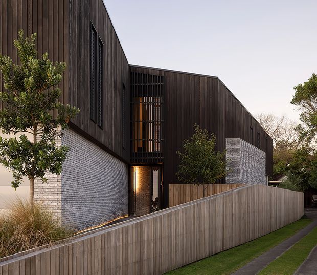 Exterior view of a contemporary multi-level residence with dark vertical timber cladding, pale grey-white brick walls, tall screened glazing, and a sloping timber boundary fence. Native-style planting and a narrow pedestrian path frame the property.