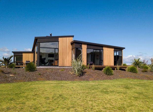 Single-storey contemporary residential building with a low, angular roofline, warm vertical timber cladding, dark metal window and door frames, and extensive floor-to-ceiling glazing. The house is set within a landscaped lawn and gravel garden with drought-tolerant ornamental planting.