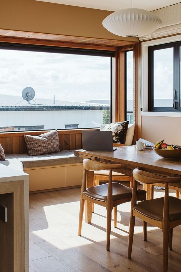 Bright open-plan dining and kitchen nook with a built-in window seat, timber-lined window reveal, large glazing, and a timber dining table with mid-century-style chairs. A sculptural white pendant light hangs overhead, while warm wood floors and joinery create a relaxed, sunlit interior. The scene includes a laptop on the table and an exterior rooftop visible through the windows.