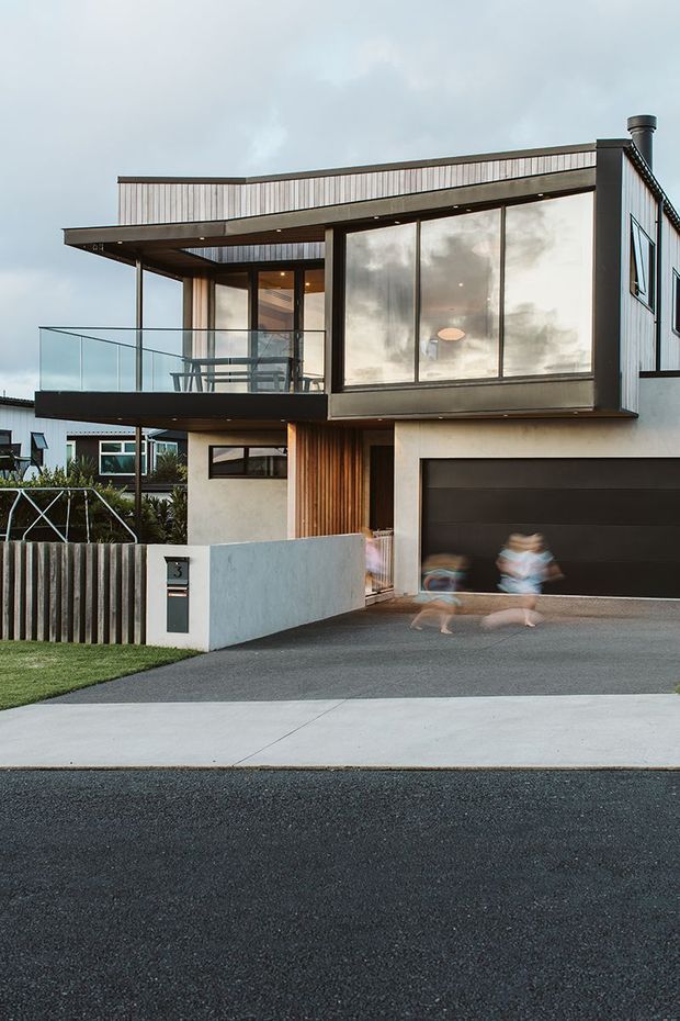 Contemporary two-storey urban residence photographed from the street, featuring a flat roof, projecting upper-level balcony with clear glass balustrade, extensive glazing, dark metal framing, pale rendered walls, vertical timber screening, and a dark double garage door. A small front lawn, concrete driveway and low boundary wall complete the frontage; blurred people are visible near the garage.