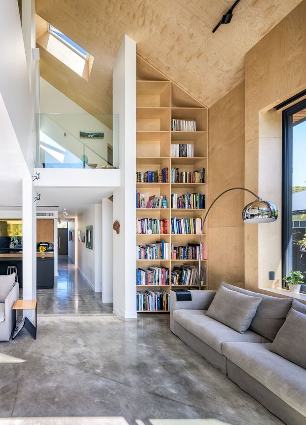 A bright, double-height living area with a large built-in plywood library and angular geometric shelving beneath a sloping plywood-clad ceiling. The room features polished concrete flooring, a grey sectional sofa, a chrome arched floor lamp, expansive glazing, and a mezzanine or upper-level walkway with glass balustrading. A hallway and glimpse of the kitchen are visible in the background.