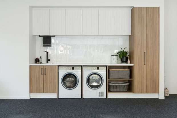A compact, highly organised laundry room with a full wall of built-in cabinetry. White overhead cupboards sit above a pale glossy tiled splashback and timber-look benchtop, with timber-look base cabinetry and a tall pantry-style cabinet framing two front-loading white washing appliances. Open shelving contains woven baskets, while a black tap, sink, plants and small accessories add contrast. The room has dark grey carpet and a clean, contemporary utilitarian feel.