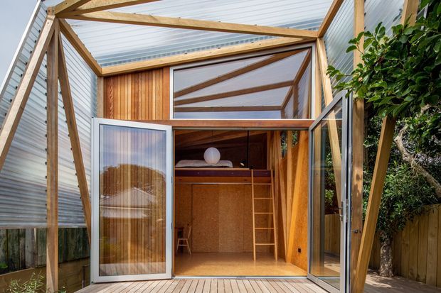 A compact timber-framed backyard retreat or studio opening onto a small timber deck. The structure features exposed diagonal bracing, translucent corrugated roofing and wall panels, warm vertical timber cladding, large glazed bi-fold doors, and a loft-like sleeping platform or storage area with a ladder inside. Dense planting and a timber boundary fence are visible around the exterior.