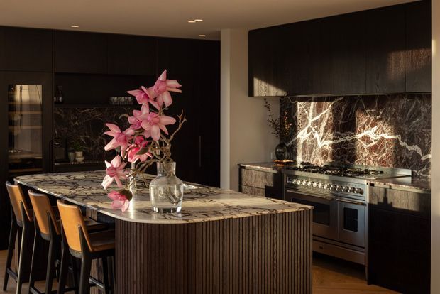 A dramatic, luxurious contemporary kitchen with a large marble waterfall island, fluted dark timber cabinetry, integrated dark wall cabinetry, and a professional stainless-steel range. A striking veined stone splashback forms the focal point behind the cooker, while warm concealed lighting, timber flooring, and black-framed details create a moody atmosphere. The island includes seating for several people and is styled with a glass vase of pink flowers.