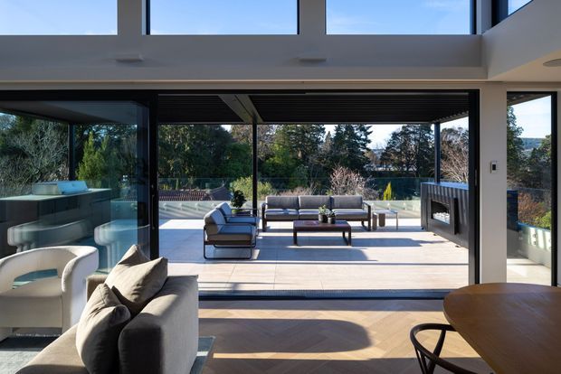 A bright, contemporary open-plan living space with expansive floor-to-ceiling glazing opening onto a covered outdoor terrace. The interior has dark timber flooring, a neutral upholstered sofa and armchair, and a rounded dining table in the foreground. Outside, a compact lounge arrangement sits on a pale tiled terrace beside a linear outdoor fireplace or privacy screen, with landscaped greenery beyond. The double-height glazing and strong indoor-outdoor connection are prominent architectural features.