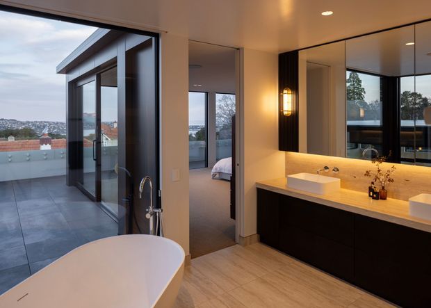 A contemporary ensuite bathroom with a freestanding white bathtub in the foreground and a wide double vanity with pale stone benchtop and dark cabinetry. Large black-framed glazing opens to a tiled terrace and provides expansive suburban views. A bedroom is directly visible through the open doorway, while a large mirrored wall, warm integrated vanity lighting, and timber-toned flooring create a refined, warm atmosphere.