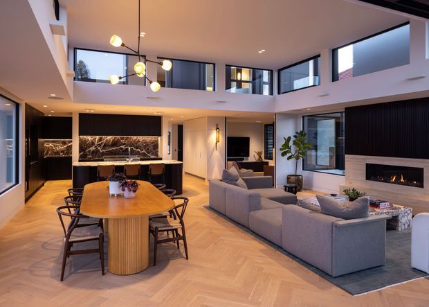 A spacious contemporary open-plan living, dining and kitchen area with a double-height void. The room features warm herringbone timber flooring, a large oval dining table, a modern kitchen with dark marble-look splashback and island, a substantial sectional sofa, black vertical wall panelling, a linear fireplace, and extensive upper-level glazing overlooking the space. A sculptural multi-globe pendant hangs above the dining area.