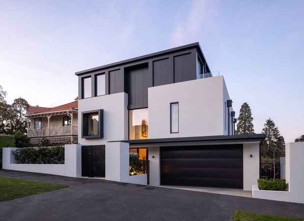 Contemporary multi-storey residential house viewed from the street at dusk. The facade combines crisp white rendered volumes with dark charcoal vertical cladding and a broad dark garage door. Narrow vertical windows, recessed glazing, a balcony railing, boundary walls, driveway and landscaped edges are visible. A traditional neighbouring house appears in the background.