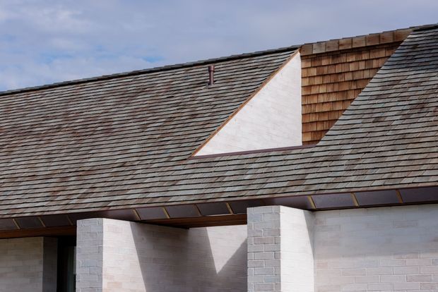 Close-up exterior view of a steeply pitched, shingle-clad roof with a prominent intersecting roof form and triangular white wall section. The facade below is composed of pale painted masonry block walls and substantial rectangular columns, with dark metal or timber detailing beneath the eaves. The image focuses on the building envelope and roof geometry against a partly cloudy sky.