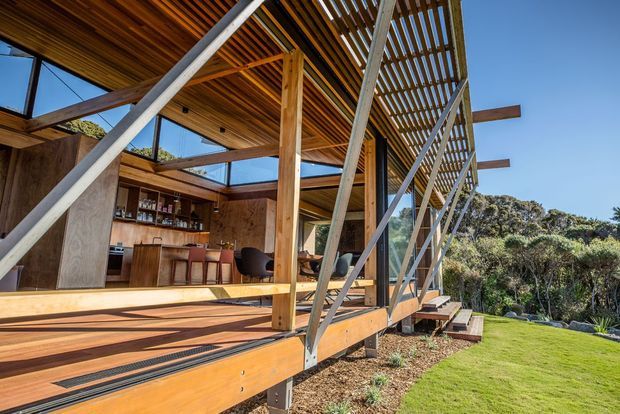 Exterior view of a contemporary timber-clad pavilion or home extension with a covered deck, open-sided slatted roof structure, extensive glazing, and a built-in bar or kitchen area visible inside. The building sits beside a landscaped lawn and dense coastal-looking vegetation, with a strong indoor-outdoor connection and exposed timber and steel framing.