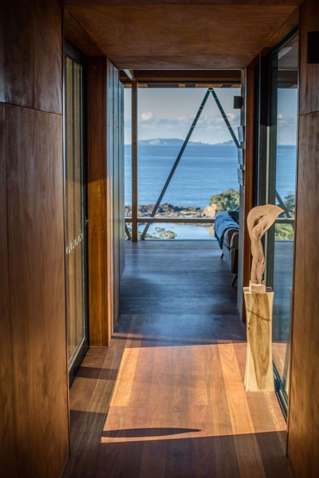 A timber-lined interior hallway leads toward a glazed bedroom or living space with expansive ocean views. Dark timber wall, ceiling and floor finishes create a warm, richly toned passage, while full-height glazing and structural framing open onto a rocky coastal setting. A sculptural timber object stands on a narrow pedestal at the right.