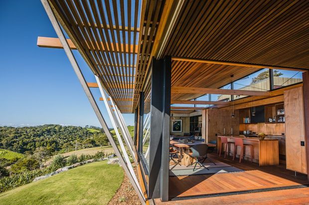 A modern timber-clad outdoor living and dining space opening onto a landscaped sloping site. The image shows extensive timber overhead louvres and soffits, exposed structural framing, dark vertical supports, large glazed openings, and a built-in timber kitchen/bar area with dining furniture. The interior connects directly to a terrace and expansive green landscape.