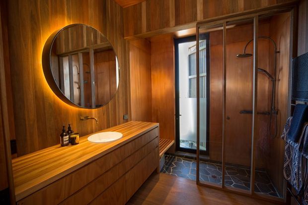 Warm, richly timber-lined bathroom featuring a floating timber vanity with a vessel basin, round backlit mirror, frameless dark-framed glass shower enclosure, wall-mounted shower fittings, and an irregular dark stone-tile floor. A glazed door brings in natural light, while integrated warm lighting enhances the intimate, luxurious atmosphere.