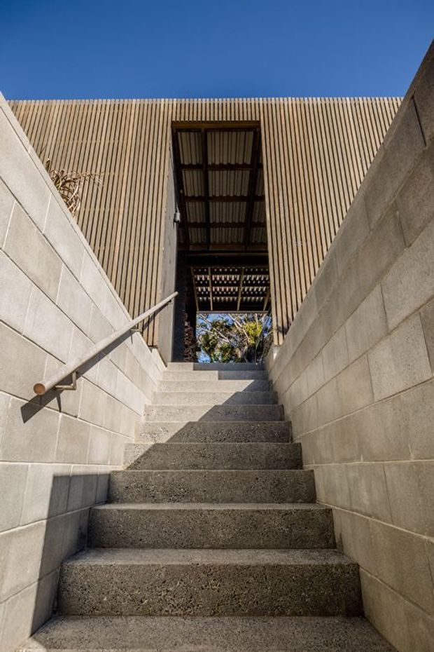 An upward-looking exterior view from a concrete stairway between grey concrete block retaining walls. A simple metal handrail runs along the left wall, leading toward a covered opening beneath a facade of closely spaced vertical timber battens. Corrugated roofing and structural framing are visible overhead, with blue sky beyond.