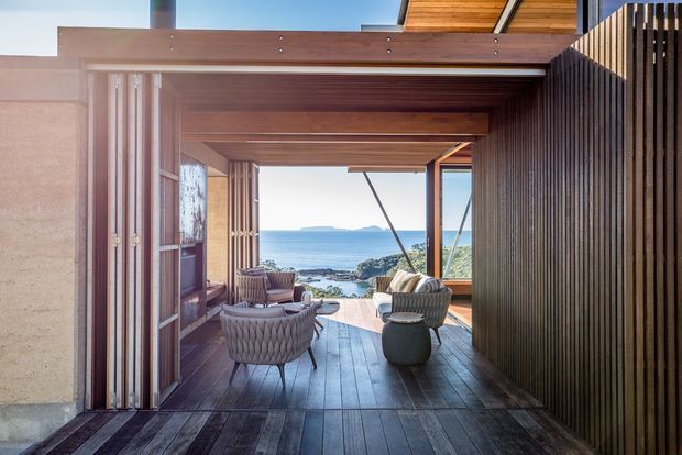 A refined coastal outdoor living area framed by timber beams and vertical slatted screening, opening toward a rocky shoreline and ocean view. The covered terrace features dark timber decking, lounge chairs, a small side table, folding timber-framed glazing, and warm wood-lined soffits.