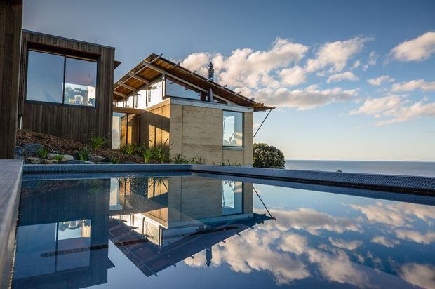 A contemporary coastal residence viewed from beside an infinity-edge swimming pool. The house features dark vertical timber cladding, exposed timber roof structure, large glazed openings, and pale stone or concrete walls. The pool reflects the building and sky, with an expansive ocean horizon beyond.