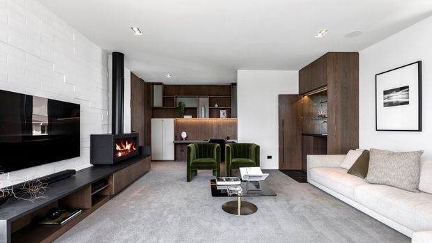 Contemporary open-plan living room with a wall-mounted television, linear fireplace, dark timber built-in cabinetry, integrated shelving and bar storage, two green lounge chairs, a white sofa, glass coffee table, and pale grey carpet. White painted brickwork and recessed ceiling lights create a clean, refined interior.