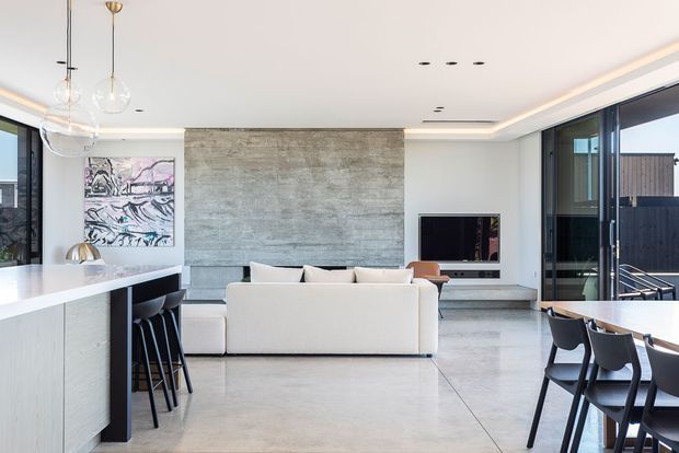 Open-plan contemporary living, dining and kitchen area with a large pale stone island, integrated dark cabinetry, a white sectional sofa, dining table, broad glazed openings and a textured concrete feature wall around the television. Minimal pendant lighting and recessed ceiling detailing create a bright, refined interior.