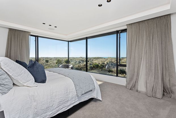 Bright contemporary bedroom with a low-profile bed dressed in white and grey bedding, full-height corner glazing, and floor-to-ceiling grey curtains. The room overlooks a natural coastal or dune landscape and has a neutral carpeted floor with a simple recessed ceiling.