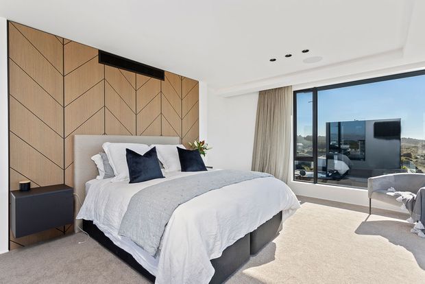 Bright contemporary bedroom with a large upholstered bed, layered white bedding and navy accent cushions. The feature wall is clad in warm timber panels arranged in a geometric chevron pattern, with integrated dark bedside storage. Full-height glazing with curtains overlooks an urban landscape, while neutral carpet and a light upholstered lounge chair create a calm, refined atmosphere.