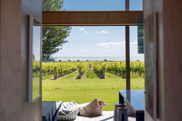 View from a timber-framed interior or covered outdoor threshold through an open glazed doorway toward neat vineyard rows and a broad distant water or horizon view. The composition is strongly axial, with timber posts and beams framing the landscape, a small deck or patio in the foreground, and a simple built-in counter or bench along the right side. Bright natural daylight and abundant greenery create a relaxed rural outlook.