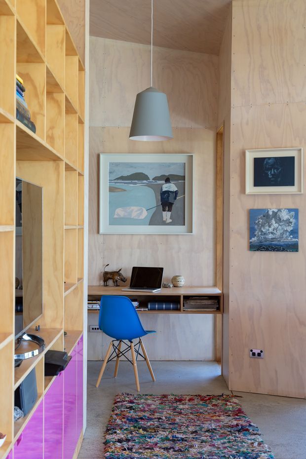 Compact plywood-lined home office nook with a built-in open shelving unit on the left, a wall-mounted timber desk, blue molded chair, pendant light, framed artwork, laptop, books and a colourful textured rug. The space has exposed plywood walls and joinery, a polished concrete floor, and a warm, playful contemporary feel.