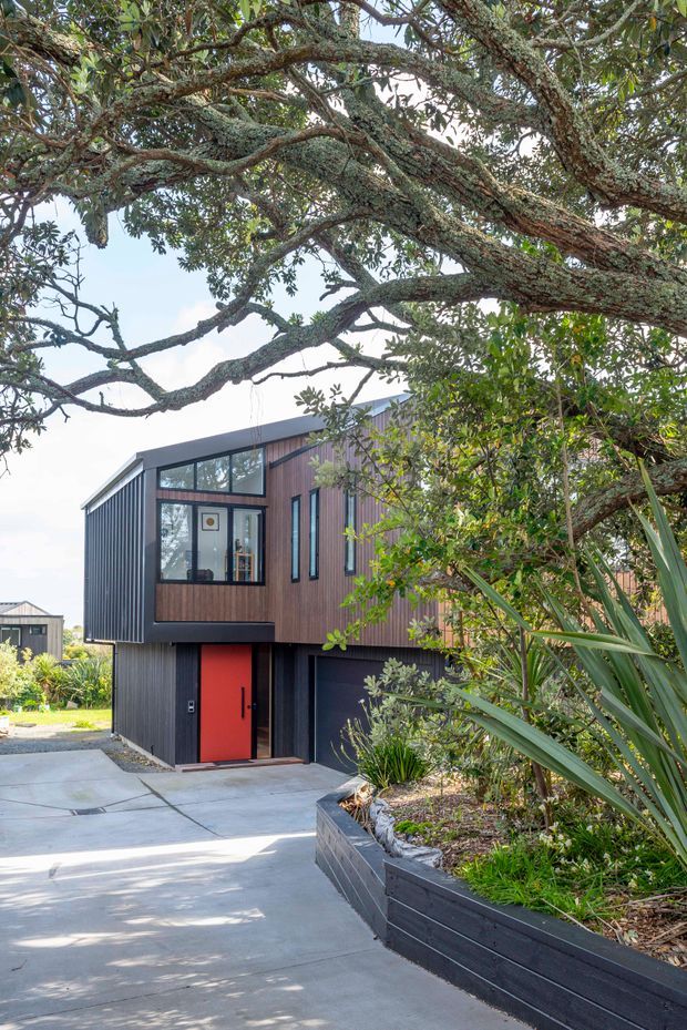 Exterior view of a compact contemporary multi-storey house framed by mature trees and landscaped planting. The building features dark vertical metal cladding, warm timber-look wall cladding, large black-framed windows, a vivid red entry door, and a concrete driveway beside raised dark garden beds.