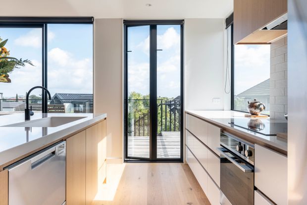 Bright contemporary kitchen with a central island, pale timber cabinetry, white stone-look benchtops, integrated stainless-steel appliances, and a white tiled splashback. Full-height black-framed glazing and a glazed door open toward an outdoor balcony or deck, bringing in abundant natural light.