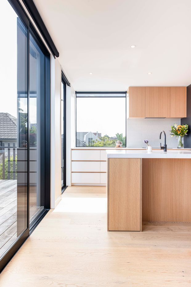 Bright contemporary kitchen with light timber cabinetry and island, white tiled splashback, pale timber flooring, and a white benchtop. Full-height dark-framed glazing opens onto a timber balcony or terrace, providing abundant natural light and suburban views. The space has clean lines, integrated appliances, recessed ceiling lights, and restrained styling.
