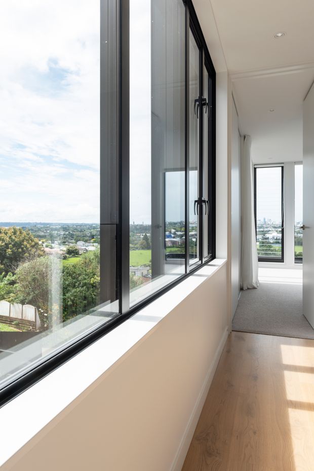 Bright, narrow residential hallway or landing with floor-to-ceiling black-framed glazing along one side, providing expansive views over a green suburban landscape. The space has white walls and ceiling with simple cornice detailing, pale timber flooring in the foreground, and a carpeted area toward the far end. Recessed ceiling lights and sheer curtains create a clean, airy contemporary interior.
