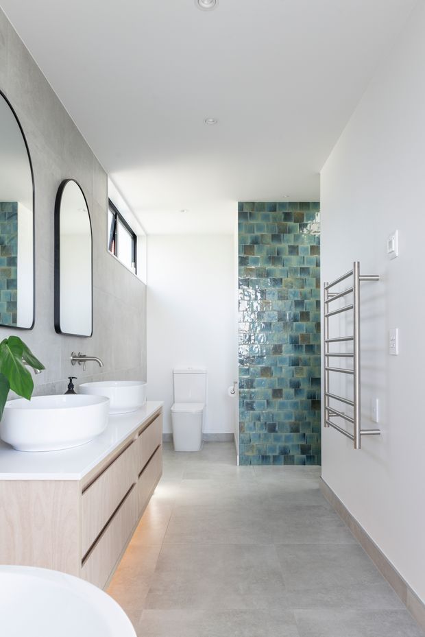 Bright contemporary bathroom with a long floating pale-wood vanity, two white vessel basins, large rounded-rectangle black-framed mirrors, a white toilet, and a stainless-steel heated towel rail. The room features a smooth grey concrete-look floor and wall finish, white ceiling and walls, and a glossy blue-green tiled shower or wet-area wall at the rear. Natural light enters through high-set windows, creating a clean and airy atmosphere.