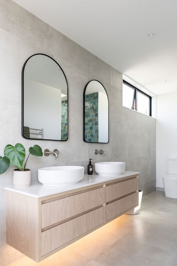 Bright contemporary bathroom featuring a floating light-wood double vanity with a white benchtop, two white vessel basins, wall-mounted brushed-metal taps, and black-framed arched mirrors. Large grey concrete-look wall and floor finishes create a clean, minimalist setting, with a high horizontal window and a glimpse of a toilet and tiled shower area. Warm under-vanity lighting adds contrast.