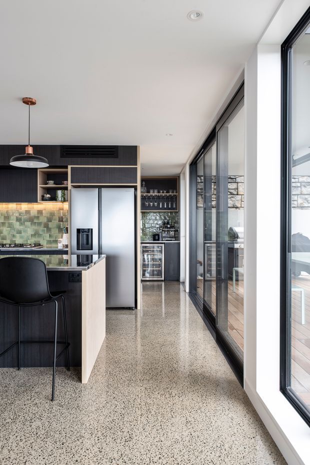 A contemporary galley-style kitchen viewed along a bright circulation zone beside expansive black-framed glazing. The kitchen features dark timber cabinetry, a pale island with black benchtop, stainless-steel refrigerator, green-toned tiled splashback, integrated wine storage, and a polished terrazzo floor. A covered outdoor deck is visible through the glazing.