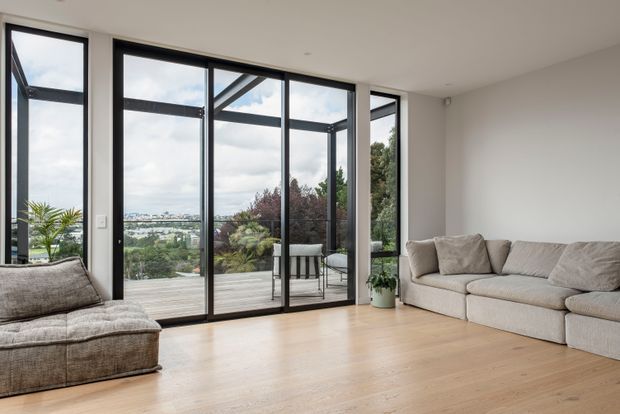 Bright contemporary living room with pale timber flooring, neutral upholstered seating, white walls, and extensive black-framed floor-to-ceiling glazing opening to a timber deck. The glazing provides views toward a landscaped suburban setting and water or distant cityscape. The room is sparsely furnished with a large sectional sofa, chaise lounge, small side table, and indoor plant.