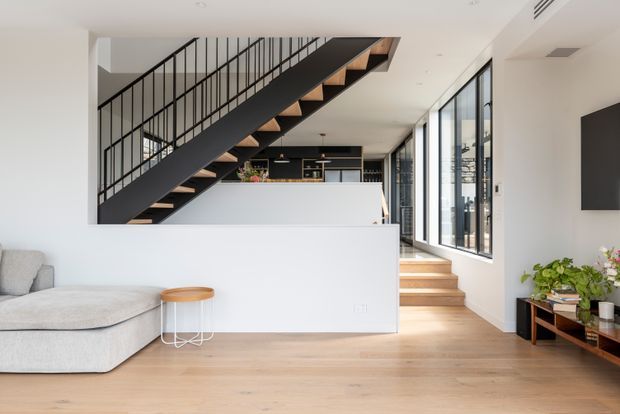 Bright contemporary open-plan interior featuring a timber staircase with black metal balustrade, light timber flooring, white walls and cabinetry, a partially visible kitchen behind a half-height partition, and a lounge area with large glazing along the side.