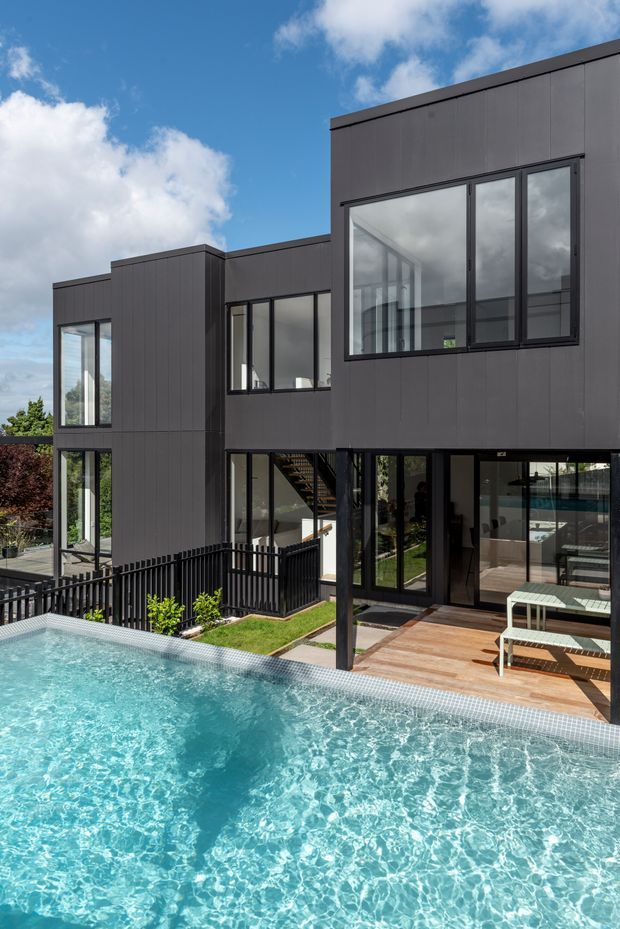A contemporary multi-level house with a dark charcoal vertical-panel facade, large black-framed windows and extensive glazing. The view looks across a bright swimming pool toward a timber pool deck and the home's glazed ground-floor living areas, with a black balustrade, landscaped garden, and blue sky visible.