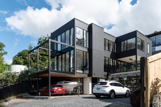 Exterior view of a contemporary multi-storey house on a sloped urban site. The home has dark vertical cladding, extensive floor-to-ceiling glazing, projecting box-like volumes, a steel-framed covered terrace or carport, a balcony, and a stone retaining wall. A gravel driveway and parked vehicles are visible in the foreground.