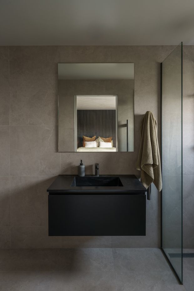 A compact, moody contemporary bathroom featuring large taupe-grey stone-look tiles on the walls and floor, a floating matte-black vanity with integrated dark basin and black tapware, and a large frameless mirror above. A frameless glass shower enclosure occupies the right side, with a heated towel rail and neutral towel beside it. The mirror reflects part of an adjacent bedroom with timber wall panelling and pillows, but the main subject is the bathroom.