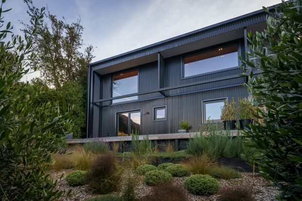 A contemporary two-storey dark-clad house viewed from a landscaped garden. The facade features vertically ribbed cladding, large rectangular windows, recessed balconies or walkways, and a raised timber deck. Native-style planting and gravel landscaping frame the building.