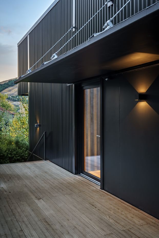 Exterior view of a contemporary dark-clad building from a timber deck. The facade features vertical ribbed metal cladding, a projecting upper balcony with metal balustrade, a timber-framed glazed door, and modern geometric wall lighting. Vegetated hills are visible in the background.